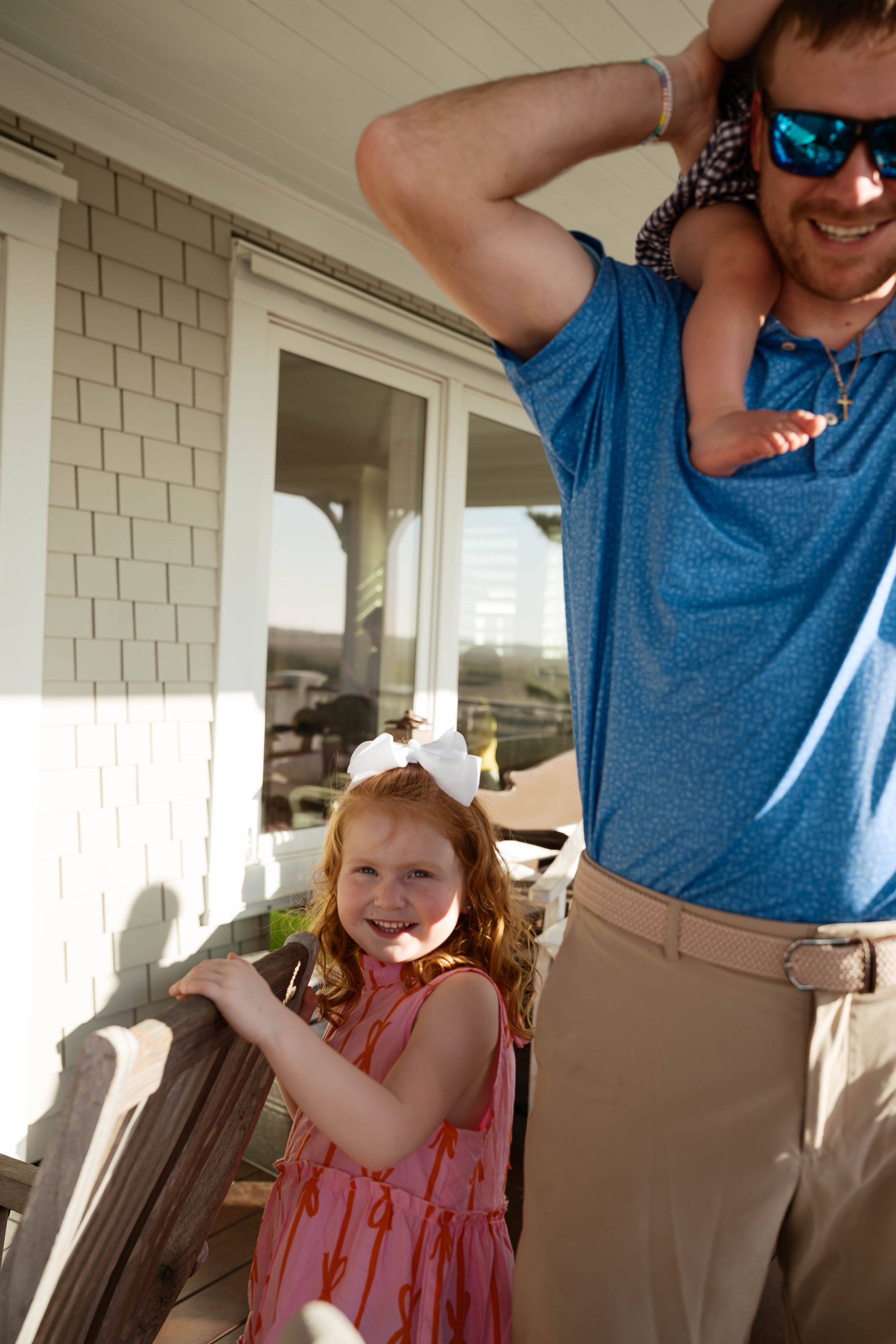 Young redhead girl in pink dress with white bow smiles near a man in blue polo carrying child on shoulders.
