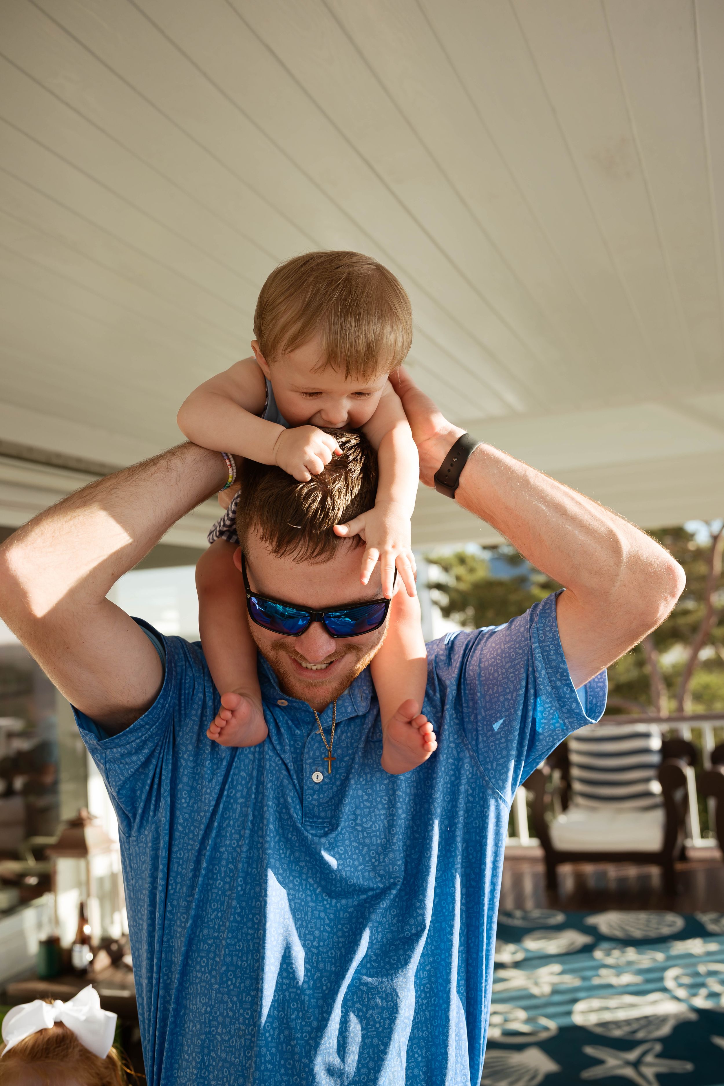 Smiling man in blue shirt and sunglasses carries a happy toddler on his shoulders under a covered patio.