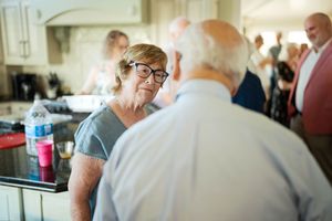 Two elderly people conversing at an indoor gathering, woman wearing glasses smiling warmly in a social setting.