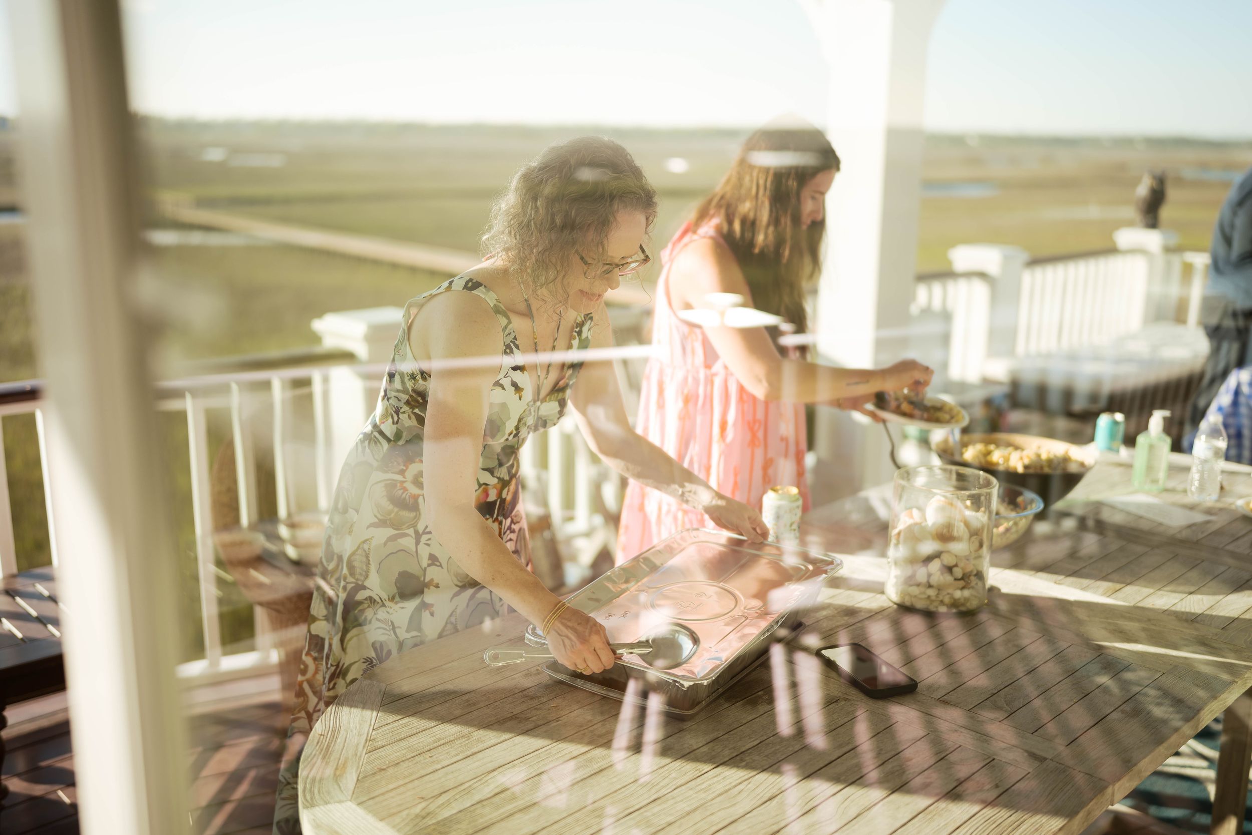 Two women enjoying a sunny outdoor deck gathering, with scenic marshland views in the background.