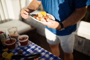 Man in blue shirt holding a plate of grilled food with lemon at an outdoor BBQ gathering with condiments on table.