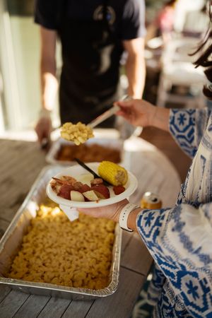 Woman serving mac and cheese onto a plate with corn, potatoes, and sausage at an outdoor buffet.