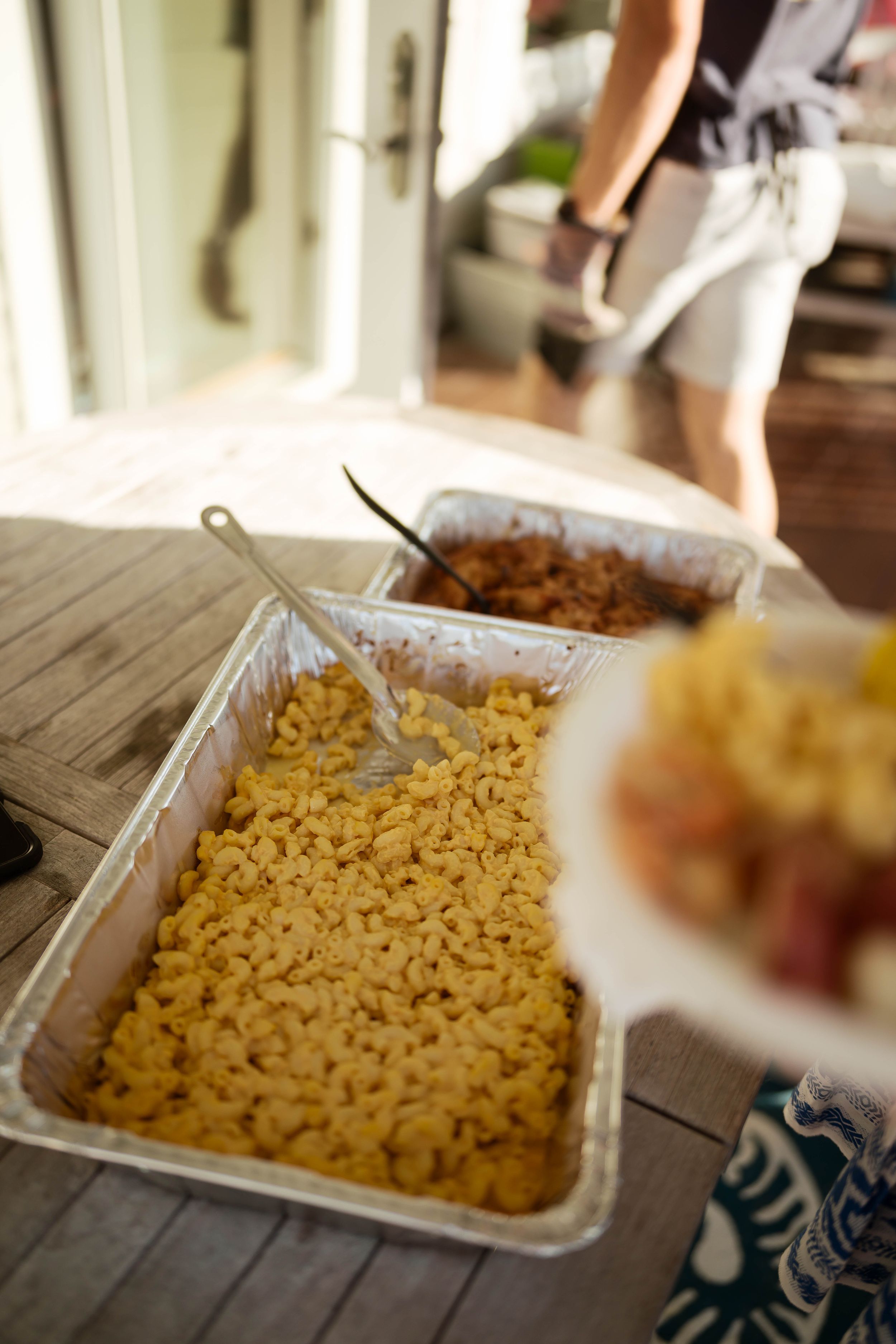 Buffet-style mac and cheese in aluminum trays on a wooden table at an outdoor gathering.