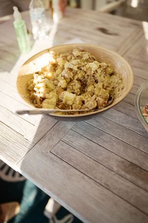 A bowl of homemade potato salad served on a rustic wooden table outdoors in natural sunlight.