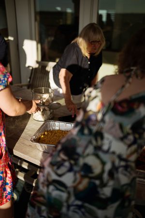 Women serving food at an outdoor gathering, with a tray of roasted corn on a wooden table in warm sunlight.