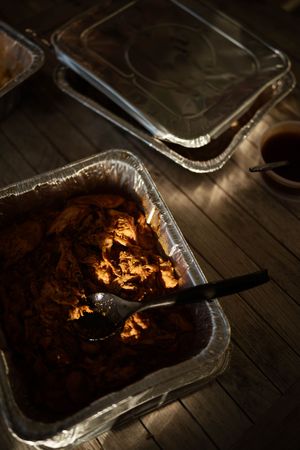 Aluminum catering tray filled with saucy pulled pork and a serving spoon, placed on a wooden table.