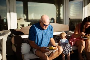 Bald man in blue shirt eating outdoors on sunny patio with woman and child enjoying food nearby.