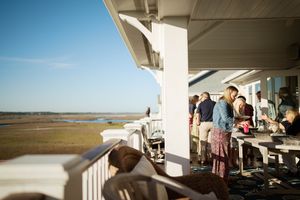 People socializing on a waterfront porch overlooking a scenic marsh estuary on a sunny day.