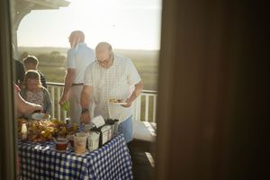 Man serving food at an outdoor buffet table with blue checkered tablecloth on a sunny deck.