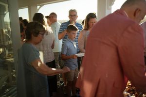 Group of people gathered indoors at a casual event, including a young boy holding a plate in warm evening light.