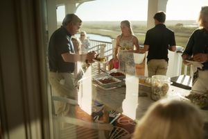 Adults socializing at a sunlit outdoor balcony party with food and drinks, overlooking a scenic waterway.