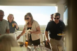 Adults socializing on a sunny beachfront deck, holding plates of food and drinks at an outdoor gathering.