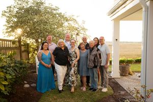 Group of ten adults smiling together outdoors near a white house with lush greenery at golden hour.