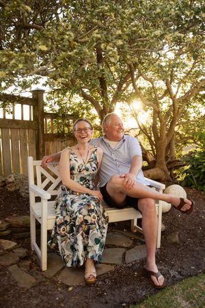 A laughing couple sits on a white garden bench surrounded by lush trees and warm sunset light.