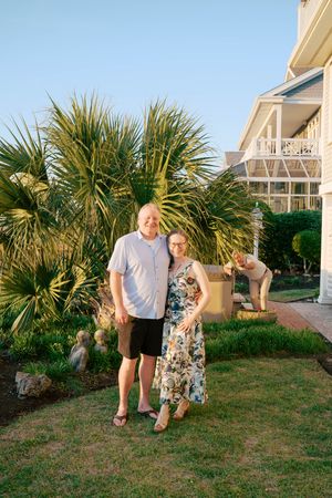 A couple smiles together in a sunny garden with palm trees beside a house.