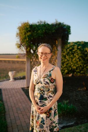 Smiling woman in floral dress and glasses stands in garden with brick path and vine-covered arbor at golden hour.