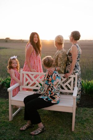 Four generations of women pose at sunset near a white bench in a field, wearing colorful dresses.