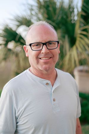 Smiling middle-aged man with black glasses and light blue henley shirt outdoors with palm trees in background.