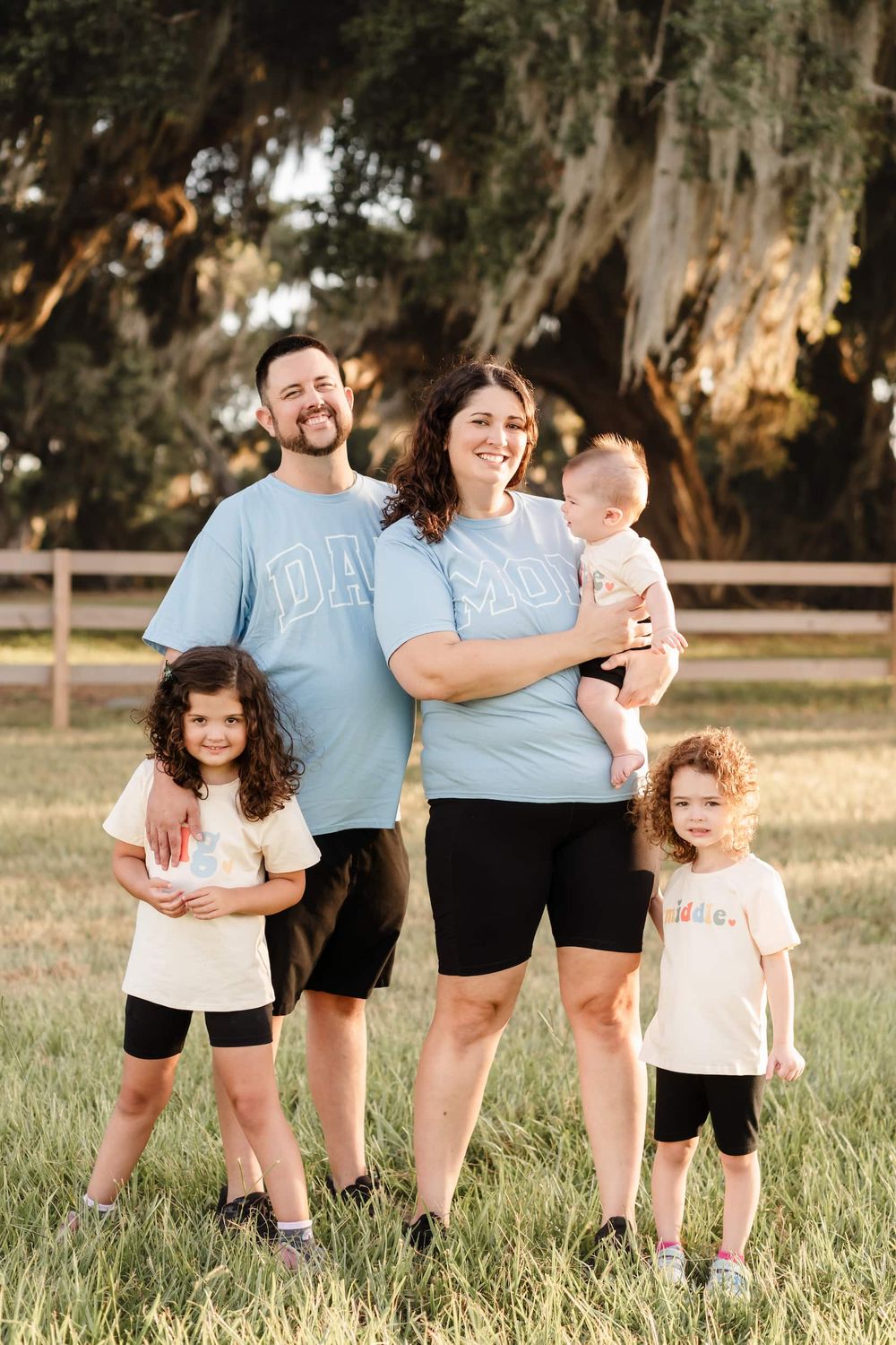 Happy family of five posing outdoors in matching light blue Dad and Mom shirts on a sunny day in a grassy field.