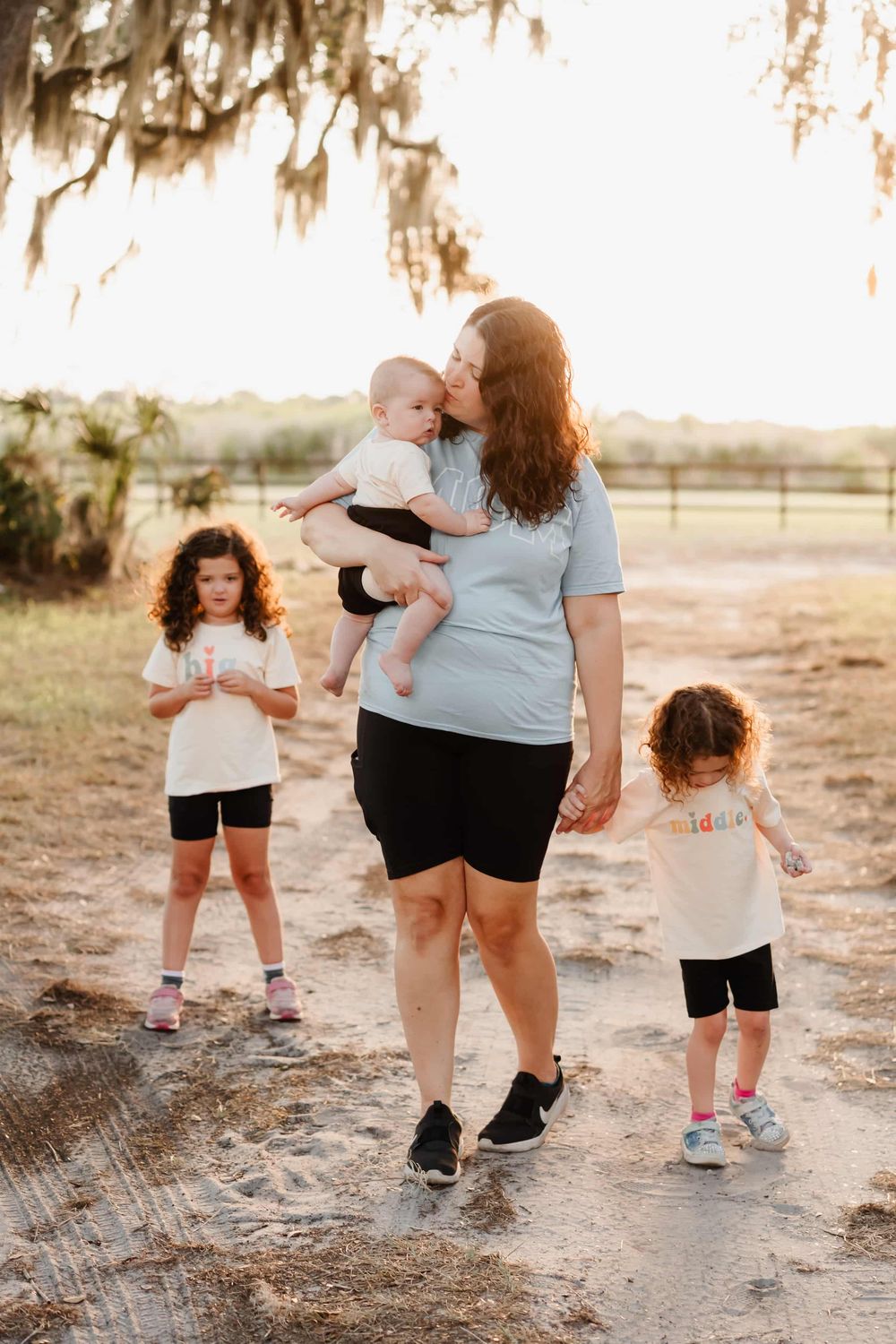 A mother kisses her baby while two young daughters walk nearby on a sunny dirt path outdoors.