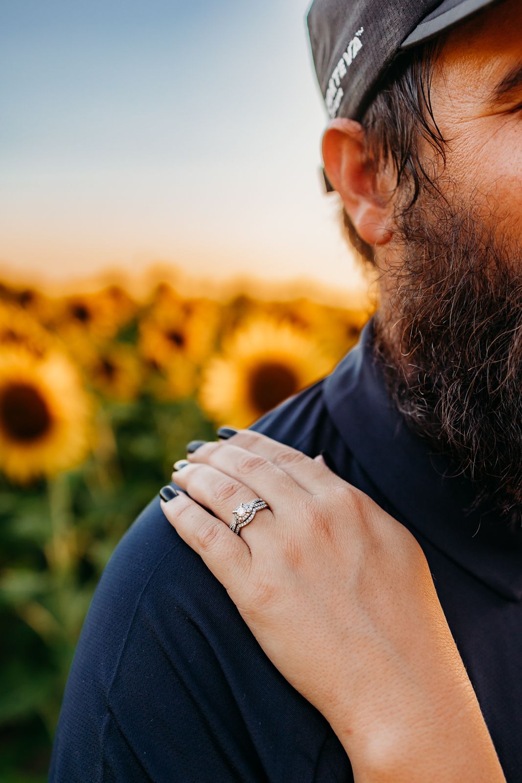 A wife's hand with her ring resting on her husband's chest in a field of sunflowers.