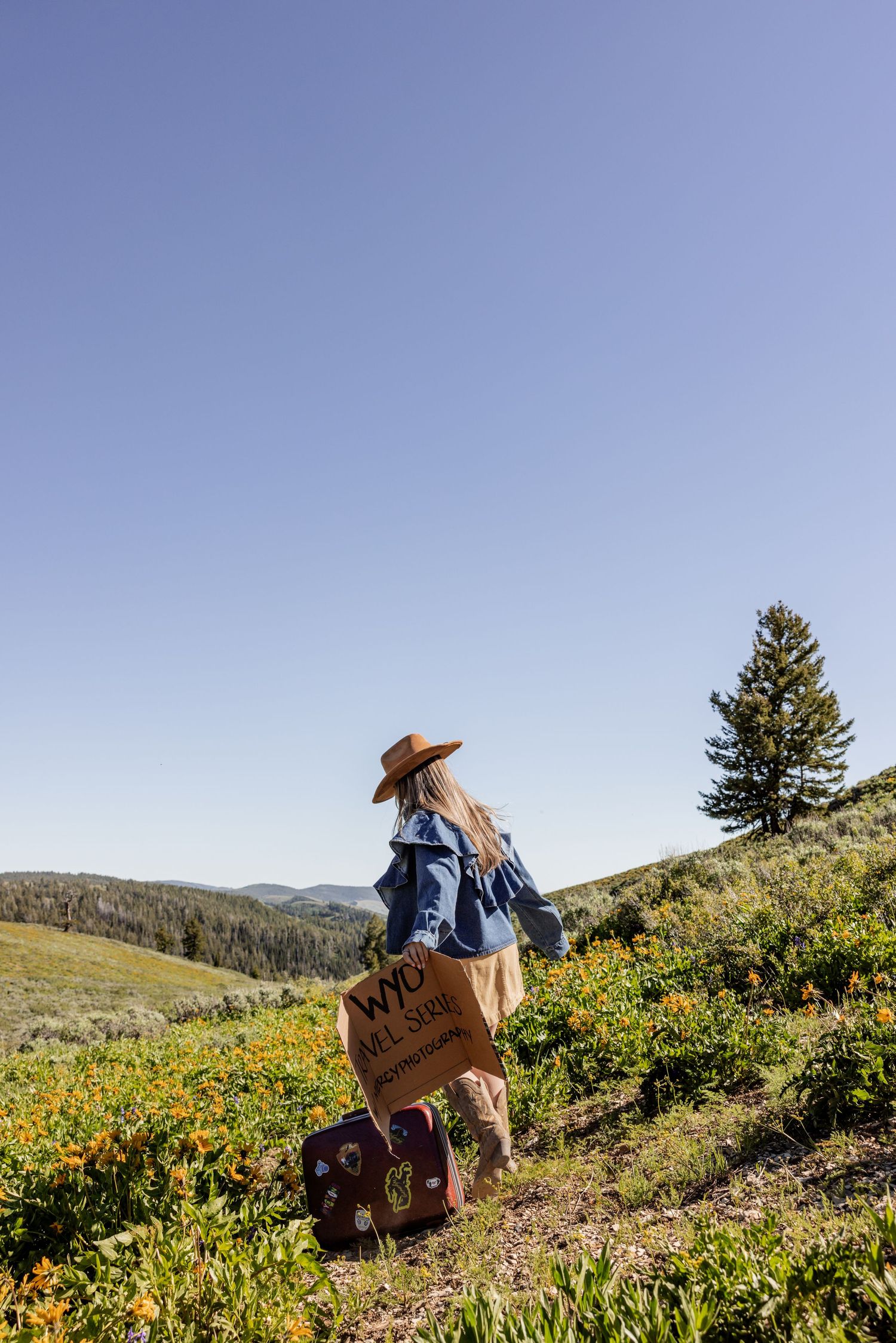 Woman in wide-brim hat and denim jacket stands on wildflower hillside with vintage suitcase and tote bag.