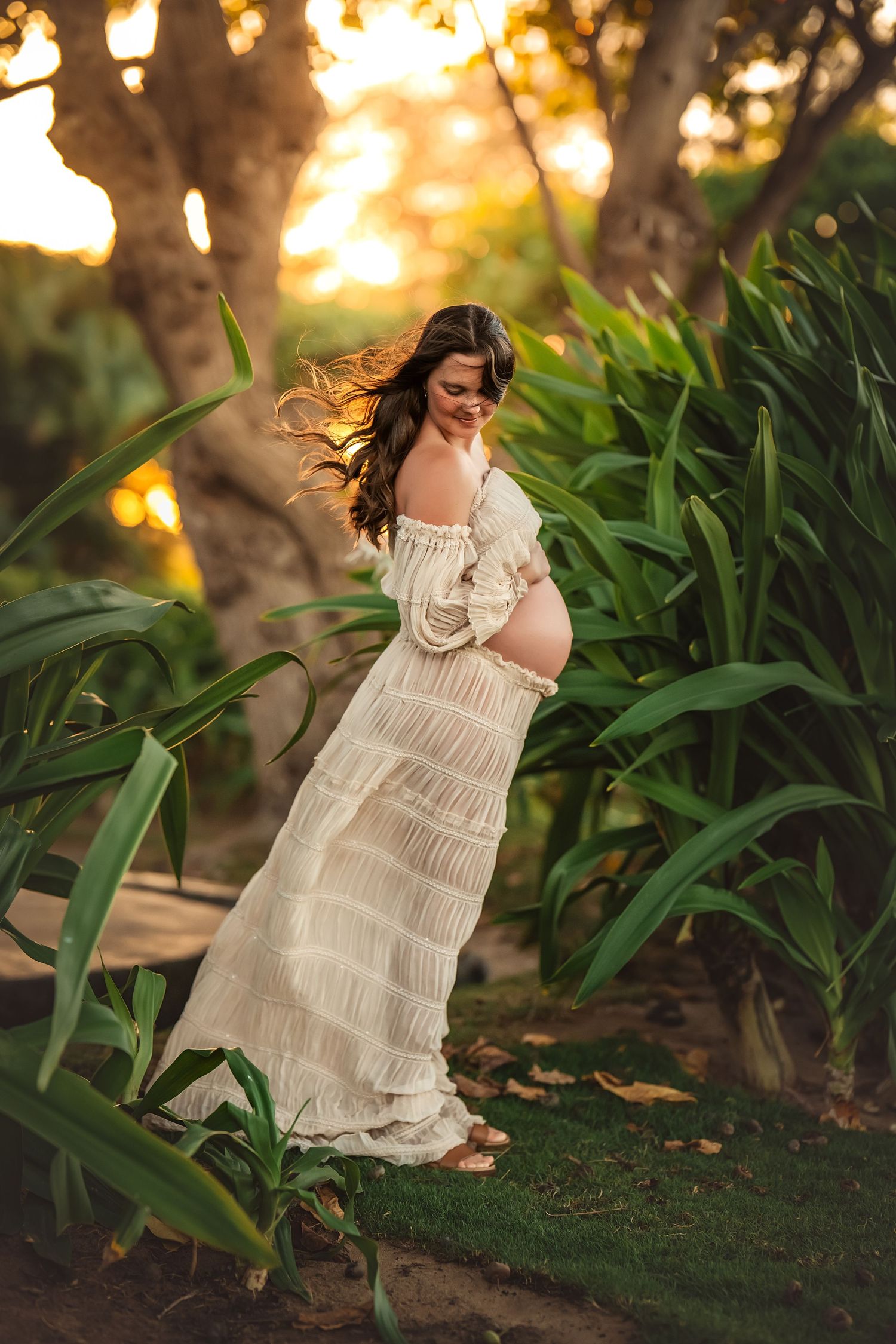 Pregnant woman in white boho dress standing among tropical plants during golden hour sunset.