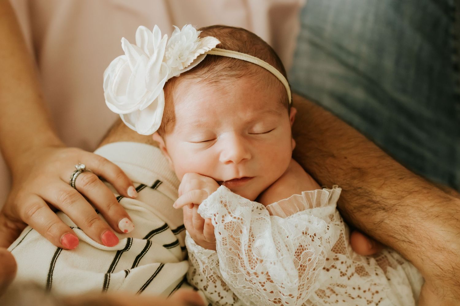 Newborn baby girl sleeping peacefully in parents arms wearing white lace outfit and floral headband.