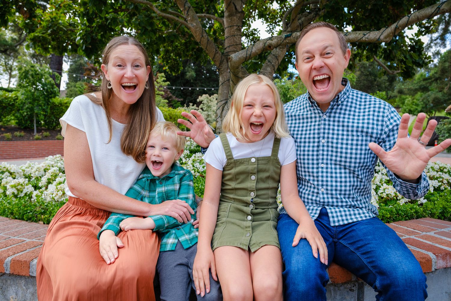 Joyful family of four laughing and making fun expressions while sitting outdoors on a bench surrounded by greenery.