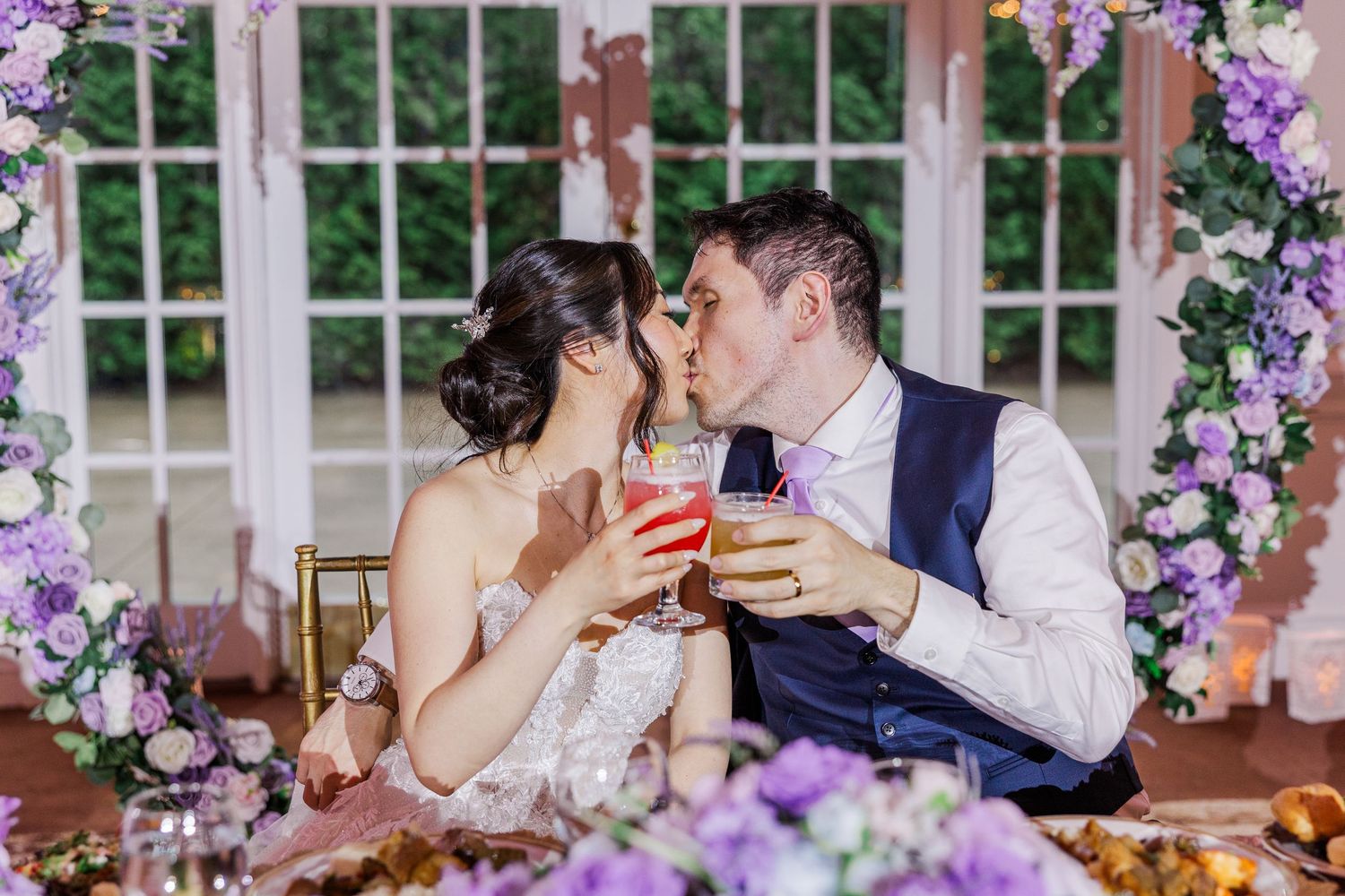 Bride and groom share a kiss while toasting with colorful cocktails at floral-decorated wedding sweetheart table.