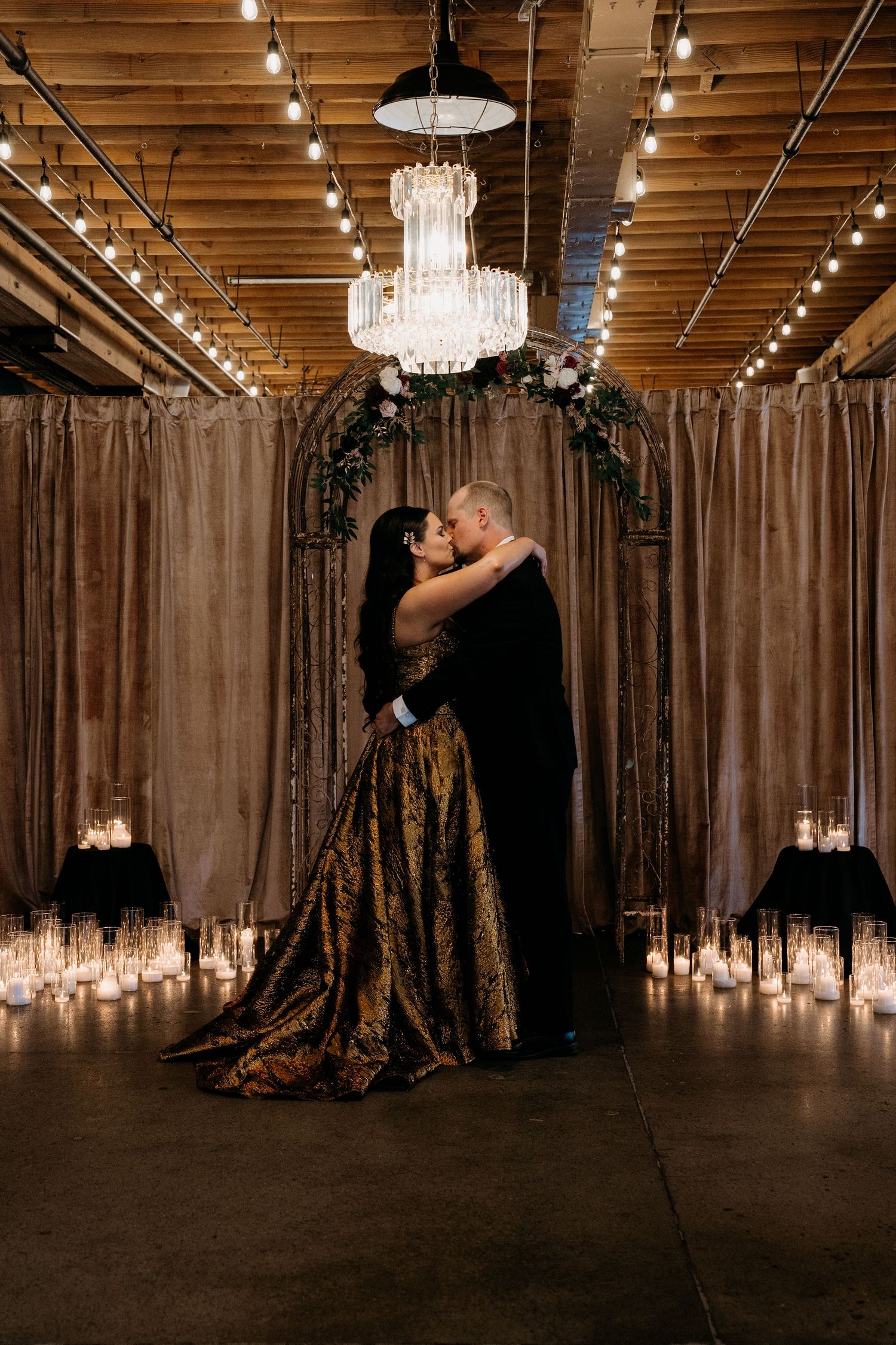 Couple sharing a kiss in golden gown under chandelier with candles and floral arch in elegant industrial venue.