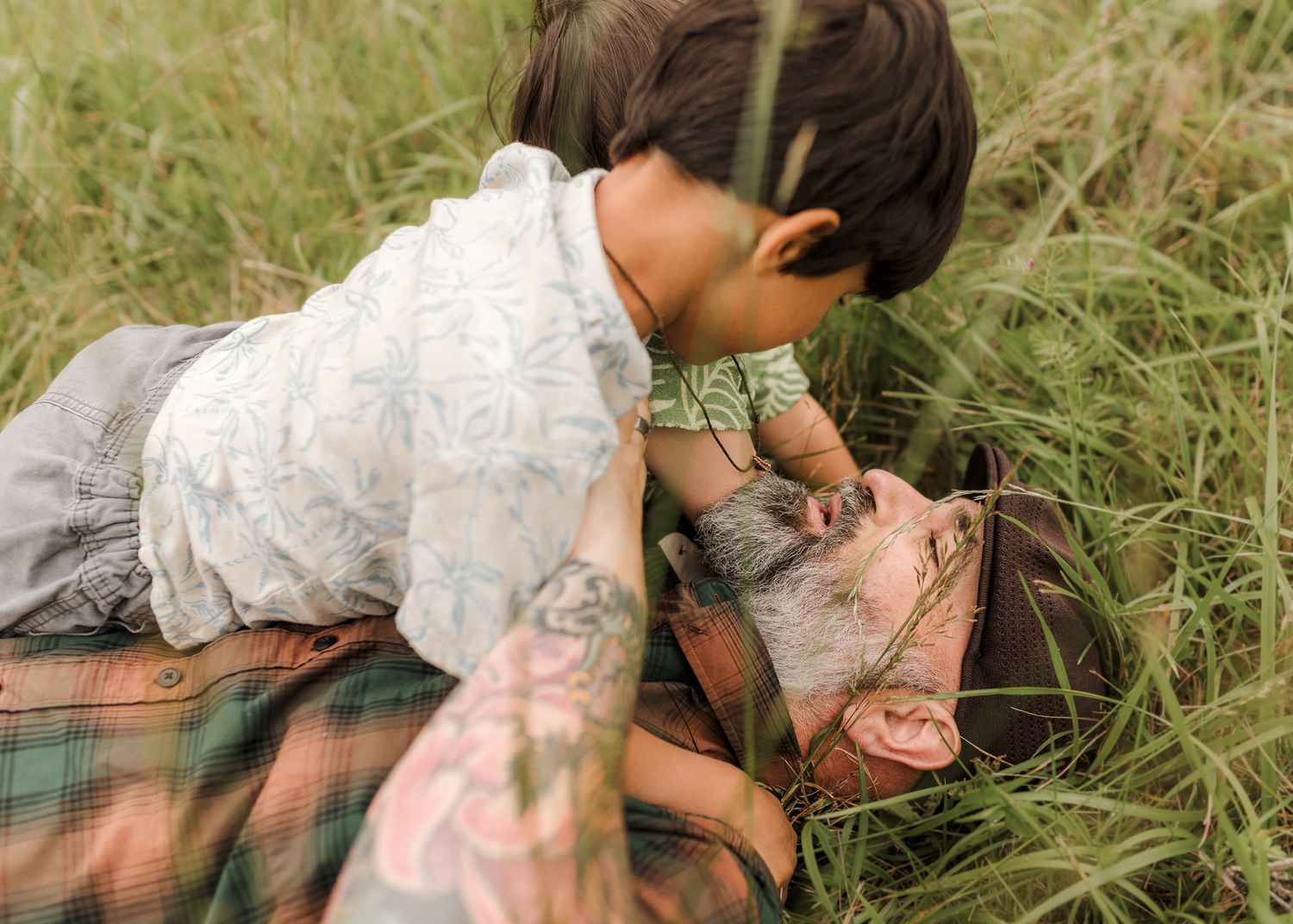 A young child leans down to kiss a tattooed man lying in tall green grass on a plaid blanket.