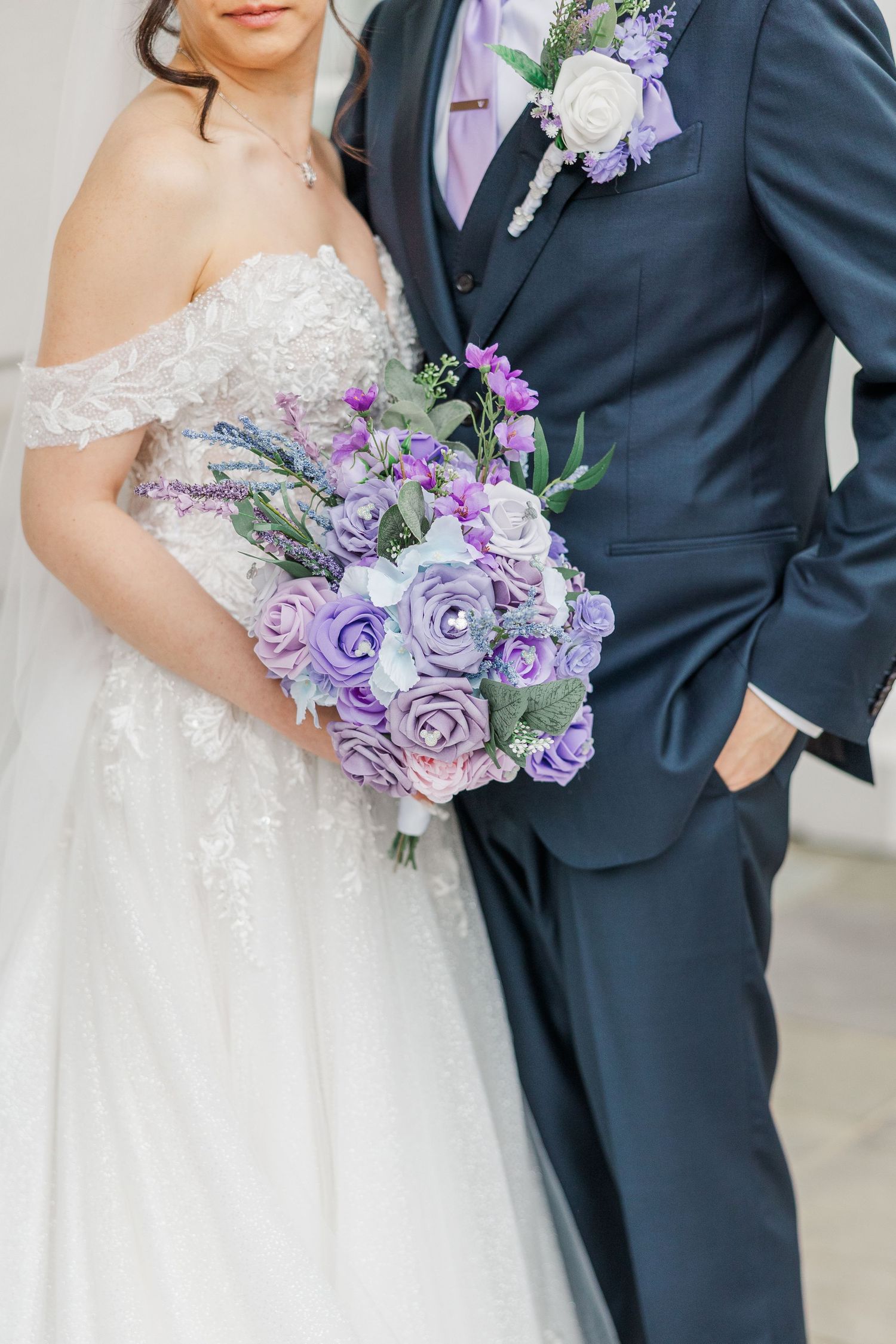 Bride in white lace gown holds purple rose bouquet beside groom in navy suit with matching boutonniere.