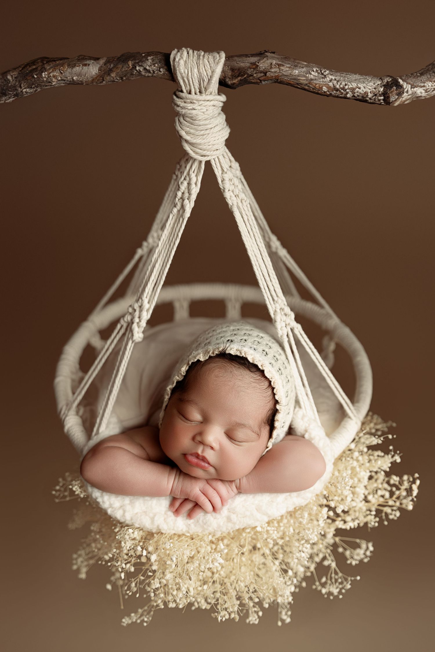 Peaceful newborn sleeping in suspended white macrame cradle with soft brown background.