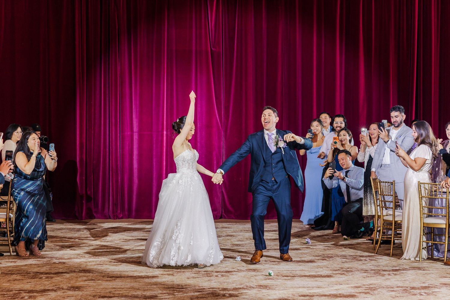 Bride and groom make grand entrance at wedding reception with red curtain backdrop, guests applauding around them.