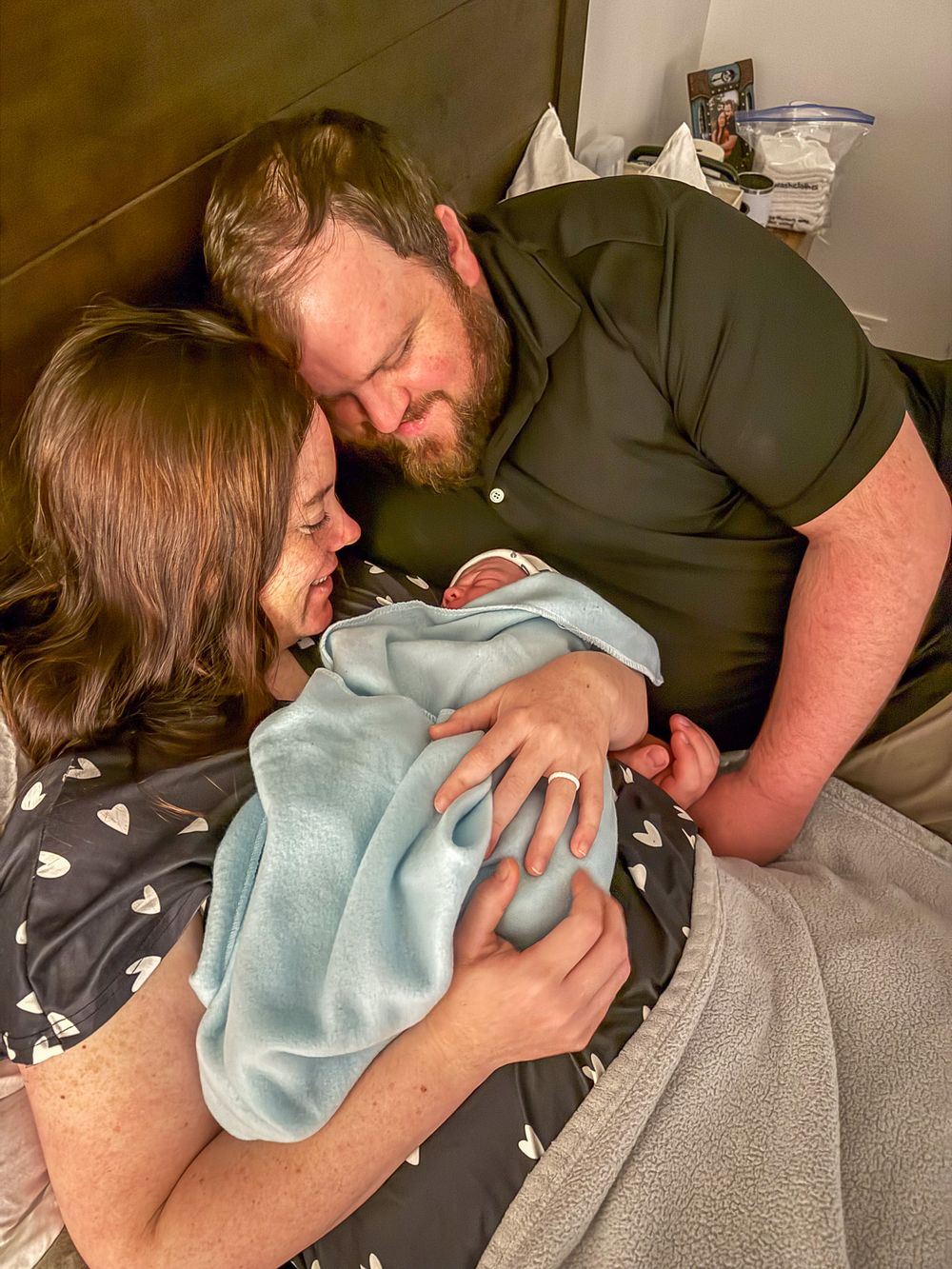 New parents cuddle their swaddled newborn in a blue blanket together on a bed.