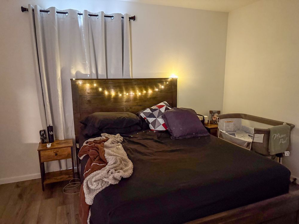 A cozy bedroom with string lights on a wooden headboard and a bassinet in corner.