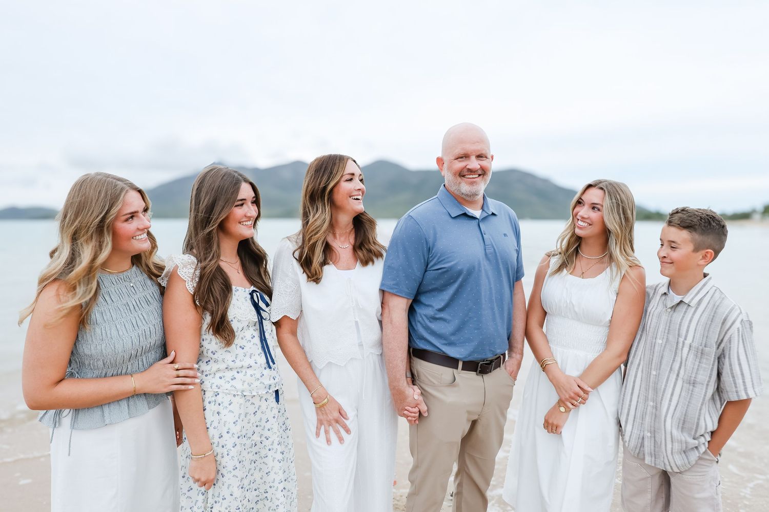 Family of six smiling together on a beach with mountains in the background on a cloudy day.