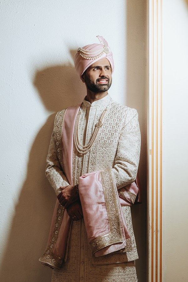 Indian groom in ornate cream sherwani with pink turban and pearl necklace posing elegantly at his wedding ceremony.
