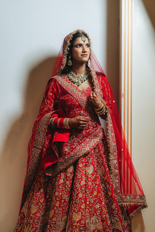 Indian bride in red embroidered lehenga with gold jewelry and red dupatta veil, posing elegantly indoors.