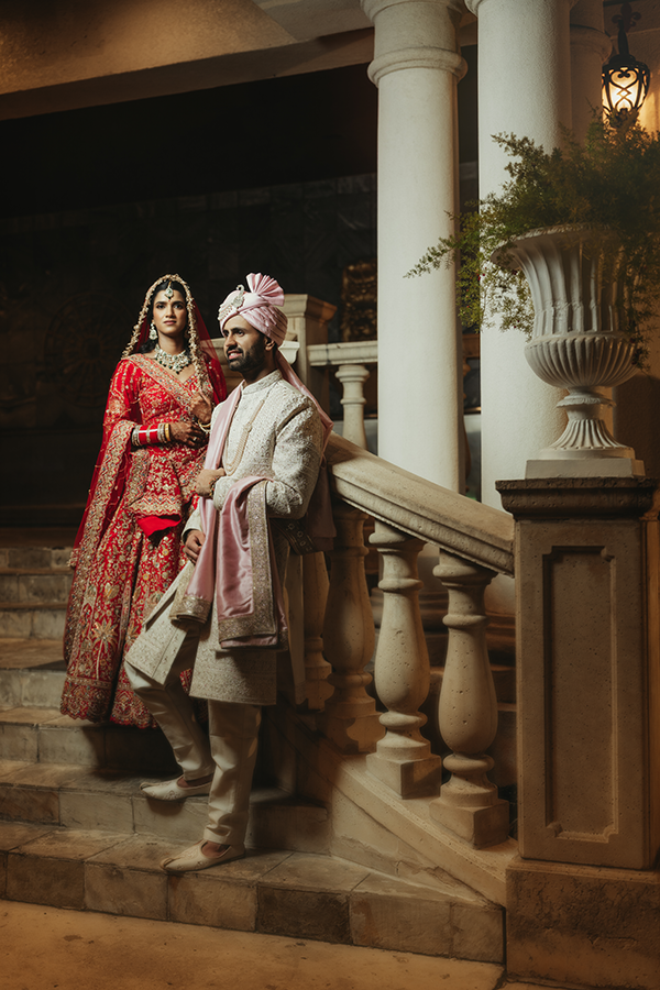 Indian bride in red lehenga and groom in ivory sherwani with pink turban pose on ornate palace steps.
