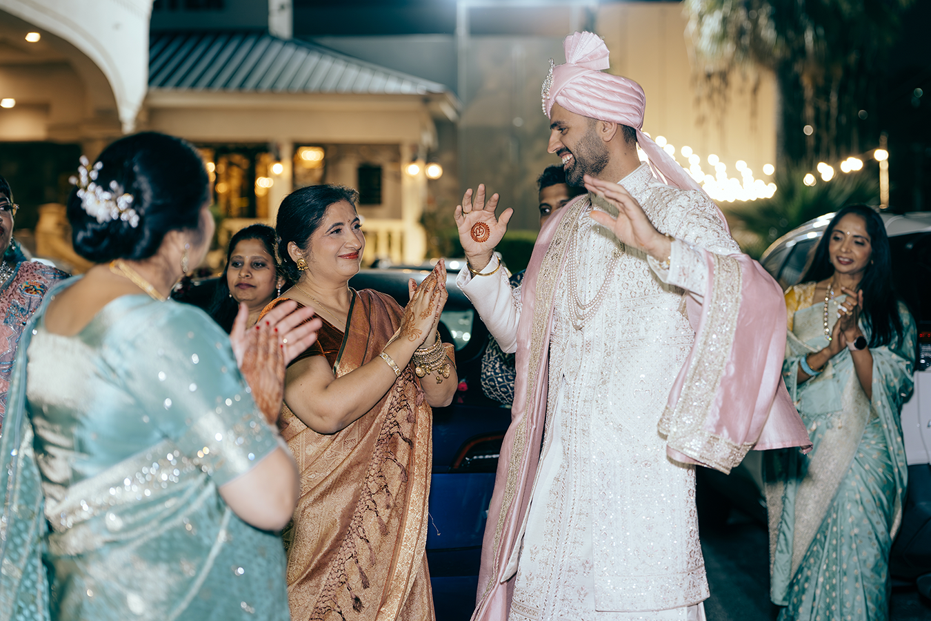 Groom in pink sherwani and turban greeted by family during Indian wedding baraat ceremony at night.