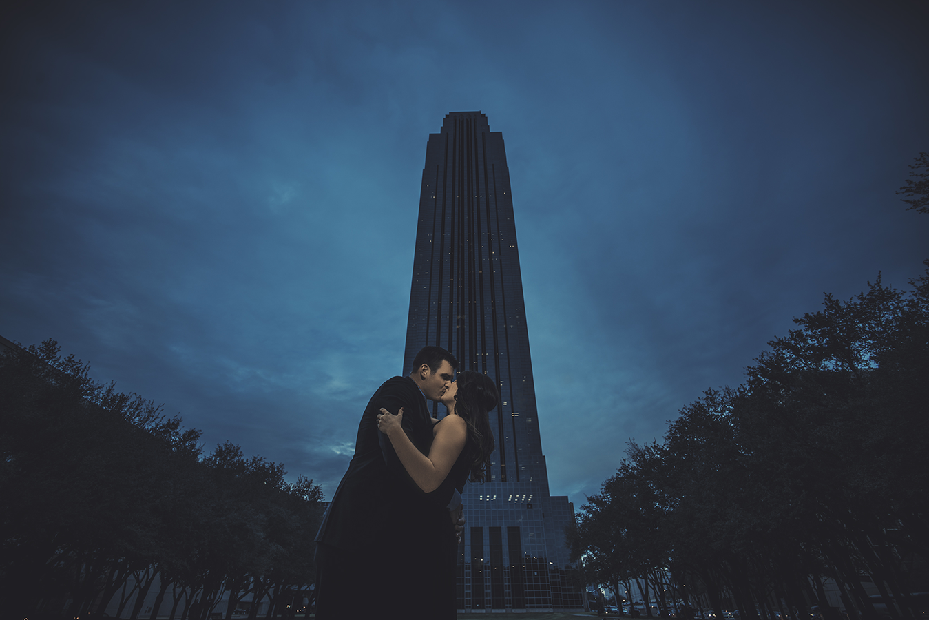 Couple embracing at dusk before a tall dark skyscraper under a dramatic cloudy blue sky.