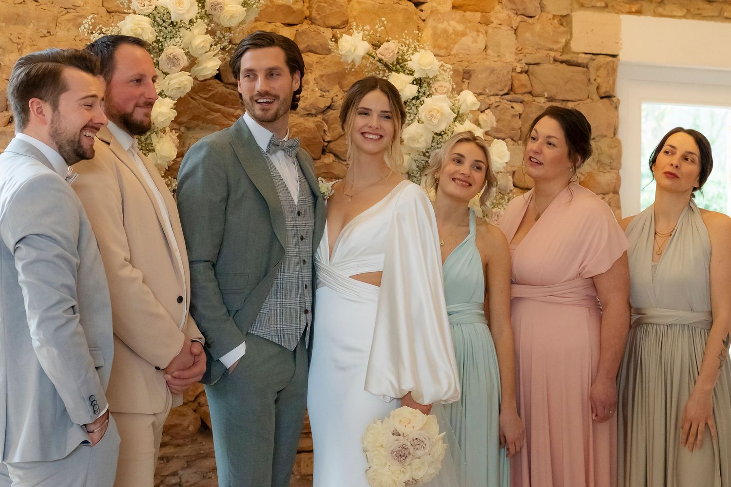 Wedding party posing together, bride in white gown with groomsmen and bridesmaids in sage and blush attire.