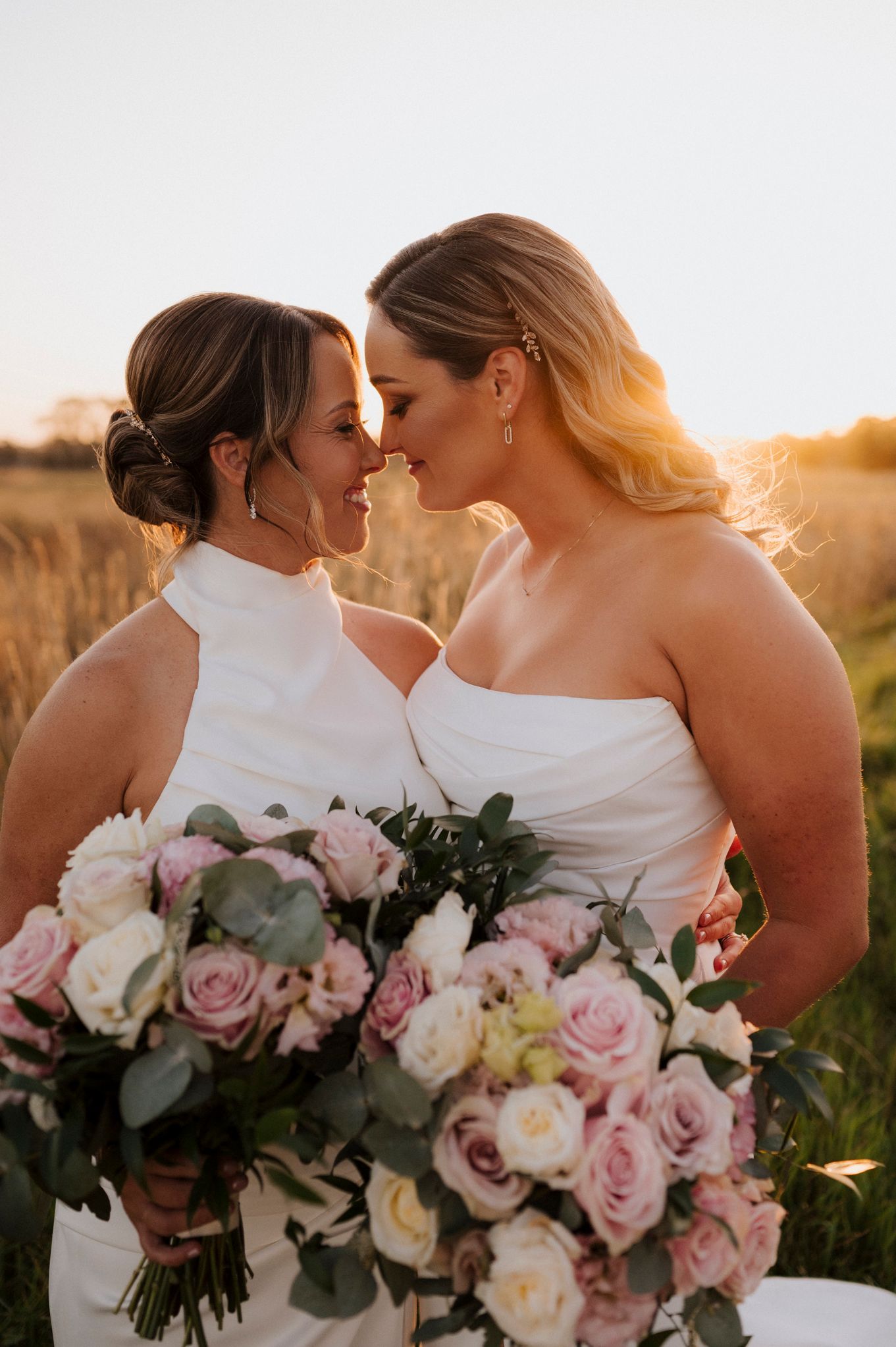 Two brides in white gowns touch foreheads at sunset, holding lush pink and white rose bouquets in a golden field.