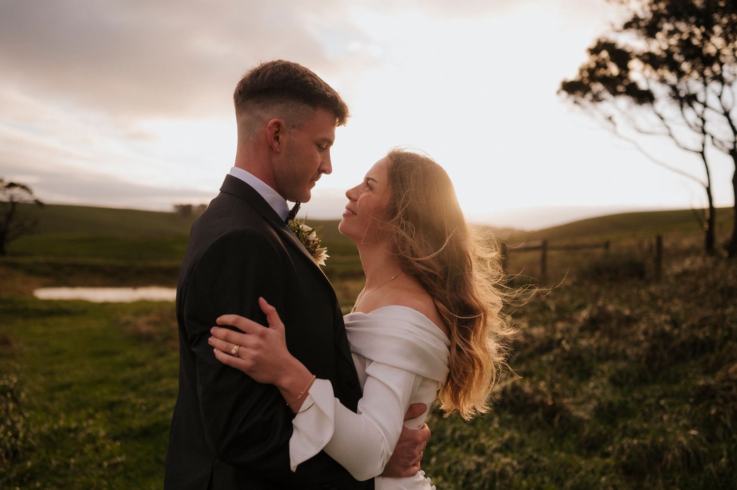 A bride and groom share a romantic embrace at sunset in a lush green countryside setting.