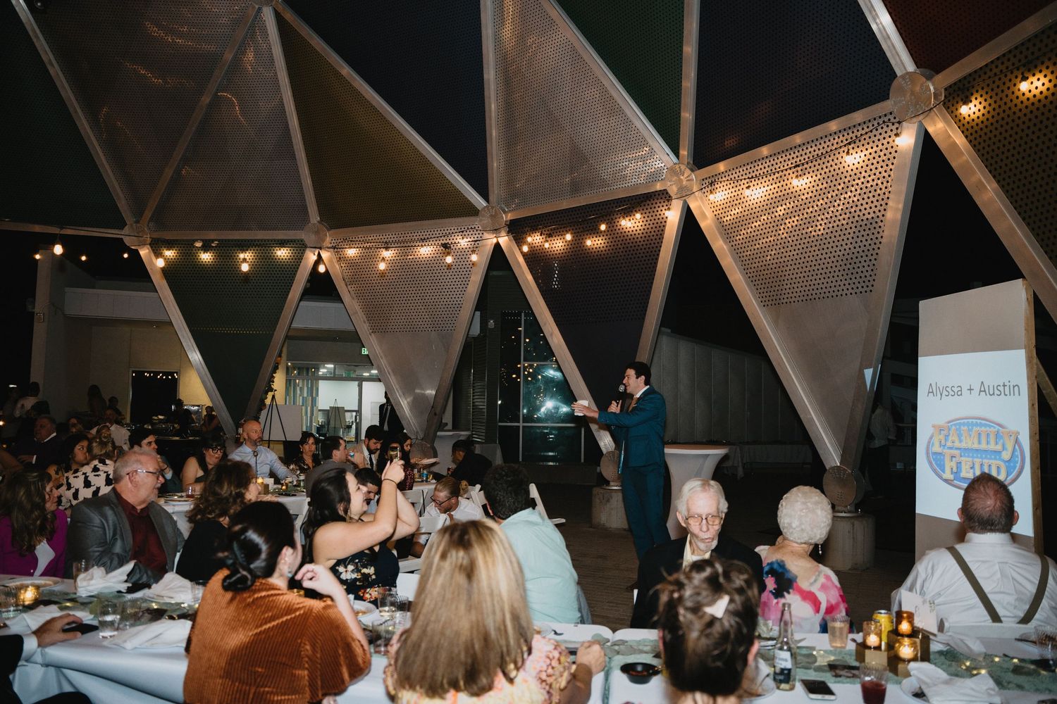A speaker addresses guests at an elegant geodesic dome dinner event with string lights overhead.