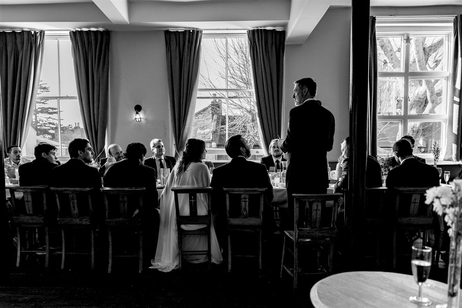 Black and white wedding reception photo showing guests seated at tables while a man gives a speech by bright windows.
