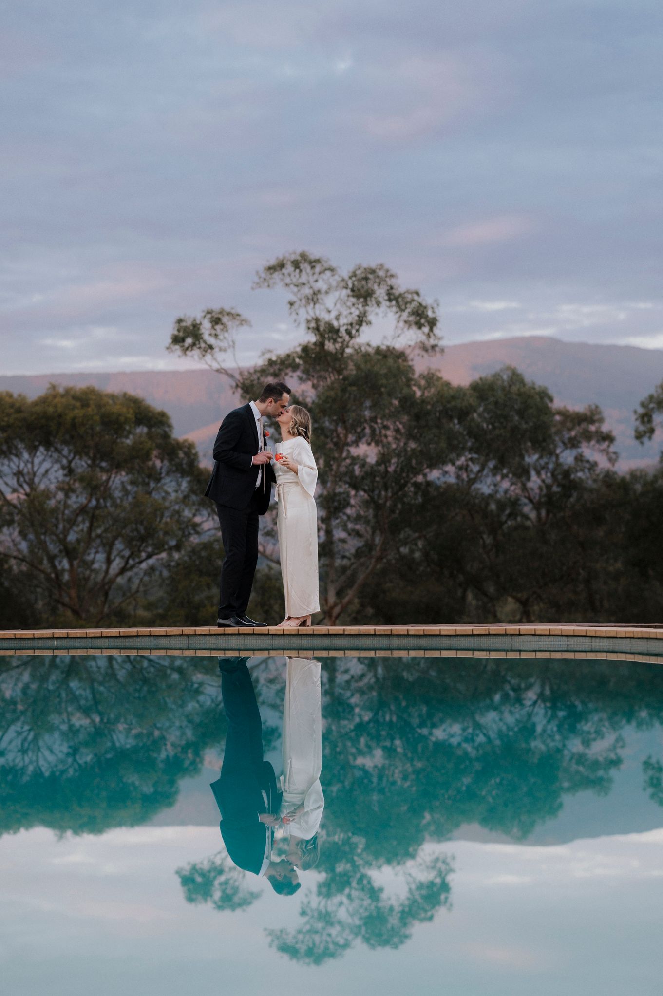 Bride and groom share a romantic kiss beside a reflective pool at dusk, with mountains and trees in the background.