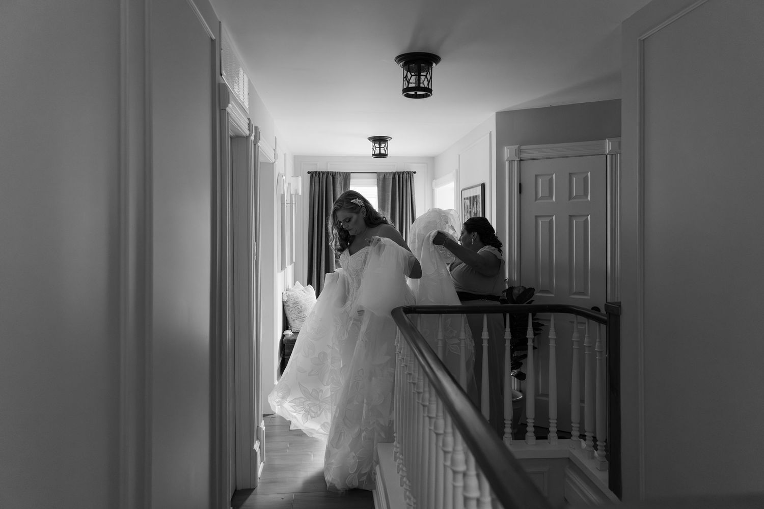 Black and white photo of a bride in a white gown standing on a balcony hallway, with a friend helping her carry her dress in Guelph, Ontario